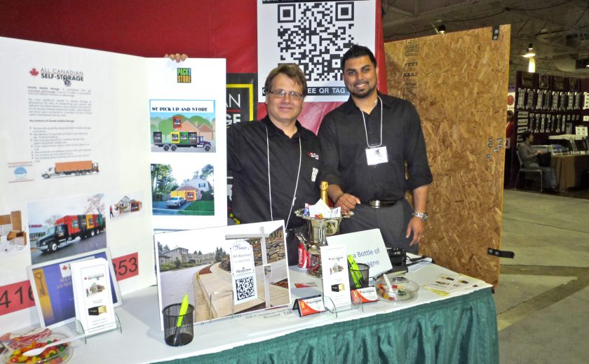 two men wearing black standing at booth, white and red boards, white table with green cloth