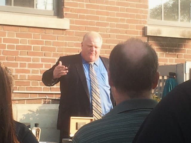 man wearing blue dress shirt and black blazer, brown brick wall with two windows, three people standing in the front