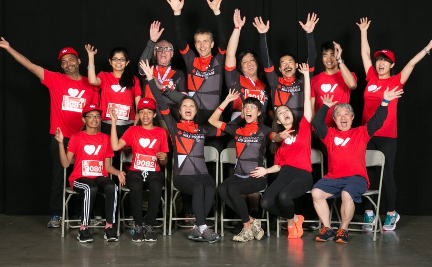 Becel Heart&Stroke Ride for Heart, large group of 15 people smiling and raising their hands up, a mix of wearing red t-shirts and a black and red biking uniform, black background